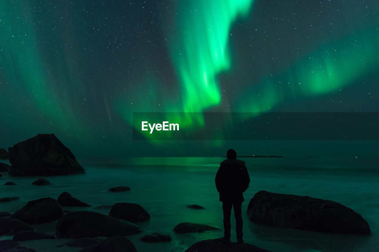 Rear view of silhouette man standing on shore against aurora borealis