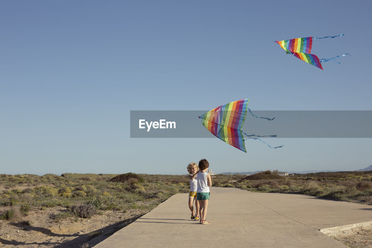 Full length of delighted little children in shorts walking on road and smiling while launching colorful kites against cloudless blue sky on sunny summer day