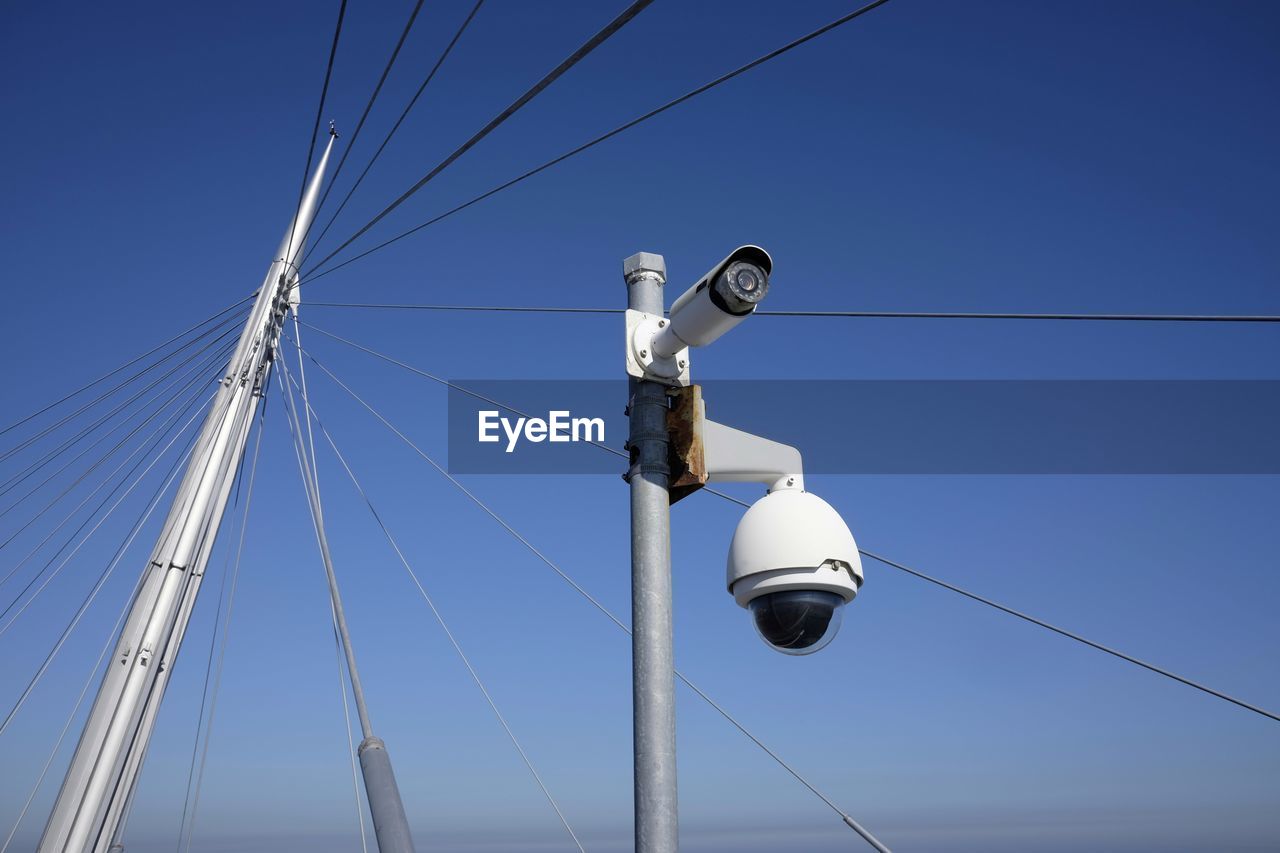 Low angle view of security camera by metallic cable against blue sky