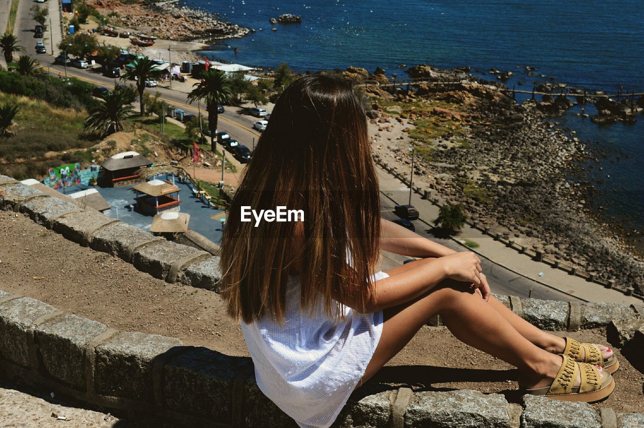 High angle view of young woman sitting on retaining wall during sunny day