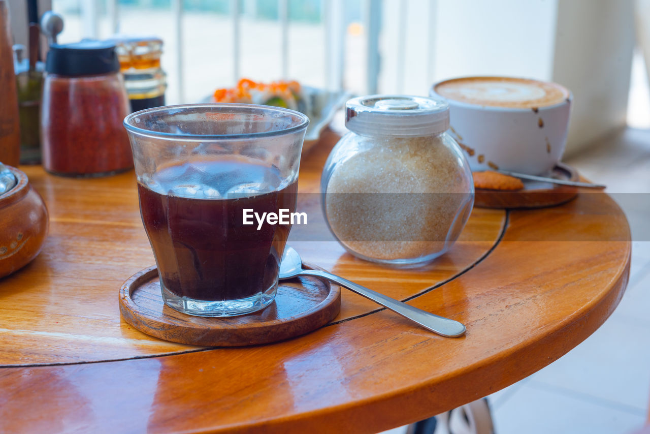 food and drink, drink, table, refreshment, indoors, household equipment, food, meal, coffee, mug, hot drink, no people, cup, glass, kitchen utensil, crockery, drinking glass, spoon, wood, cafe, day, freshness, still life, tea, breakfast, saucer, focus on foreground, eating utensil, coffee cup
