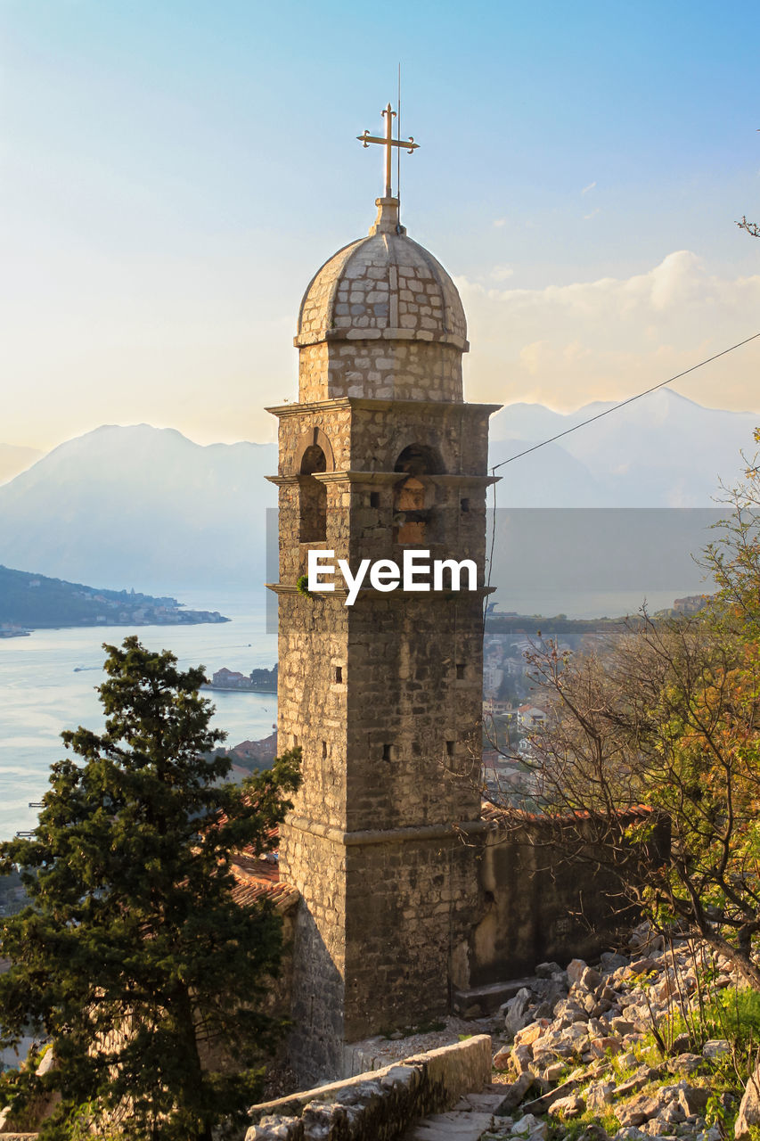A christian stone tower with a cross on its dome against a background of sea and mountains