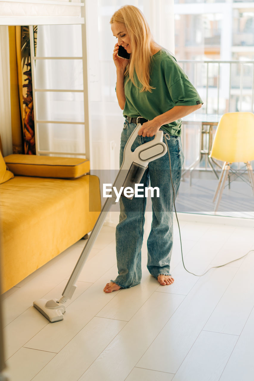 A woman talking on a mobile phone vacuums the floor in the room.