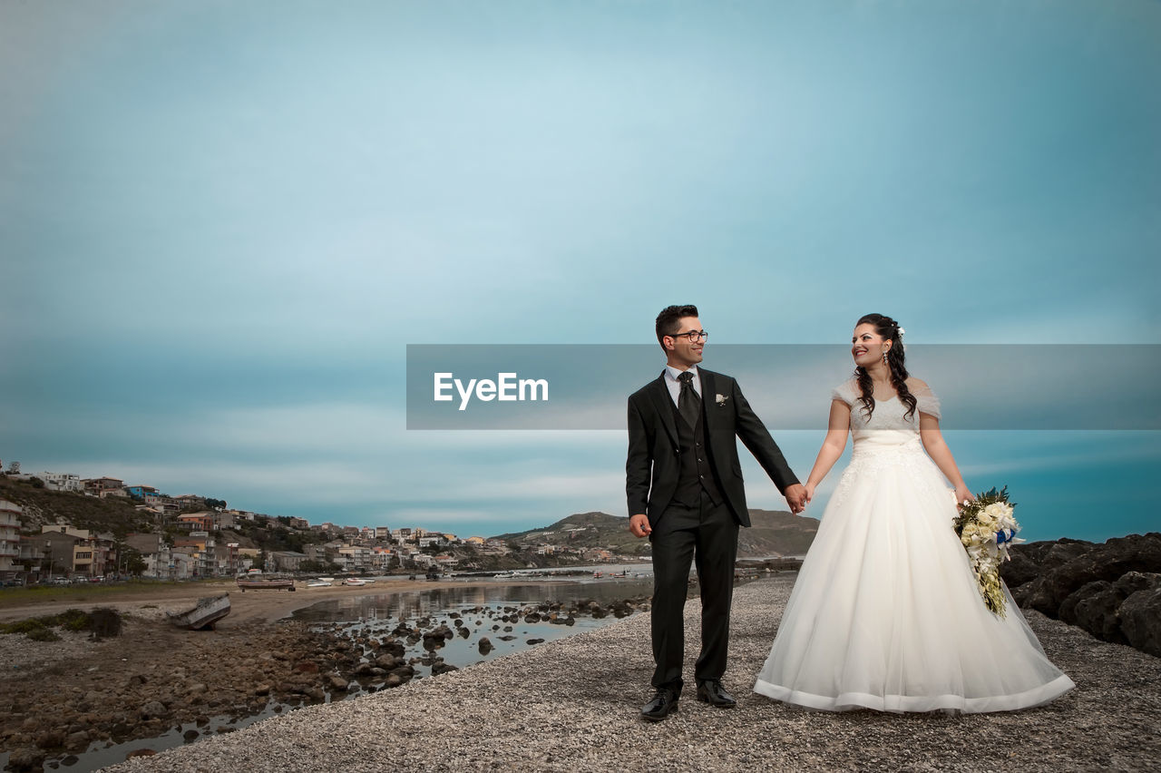 Wedding couple holding hands while standing against cloudy sky