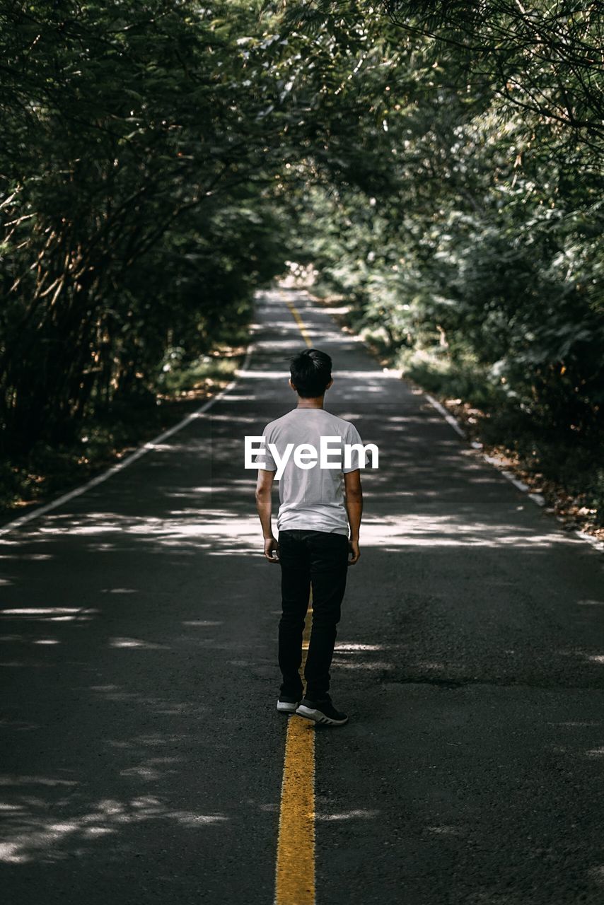 REAR VIEW OF MAN WALKING ON ROAD ALONG TREES