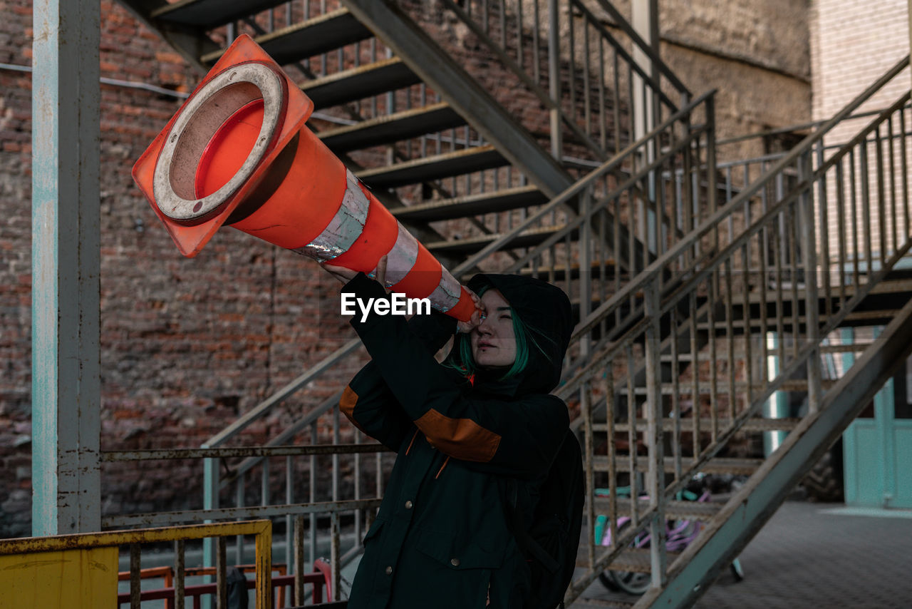 Side view of young woman looking through traffic cone while standing against building