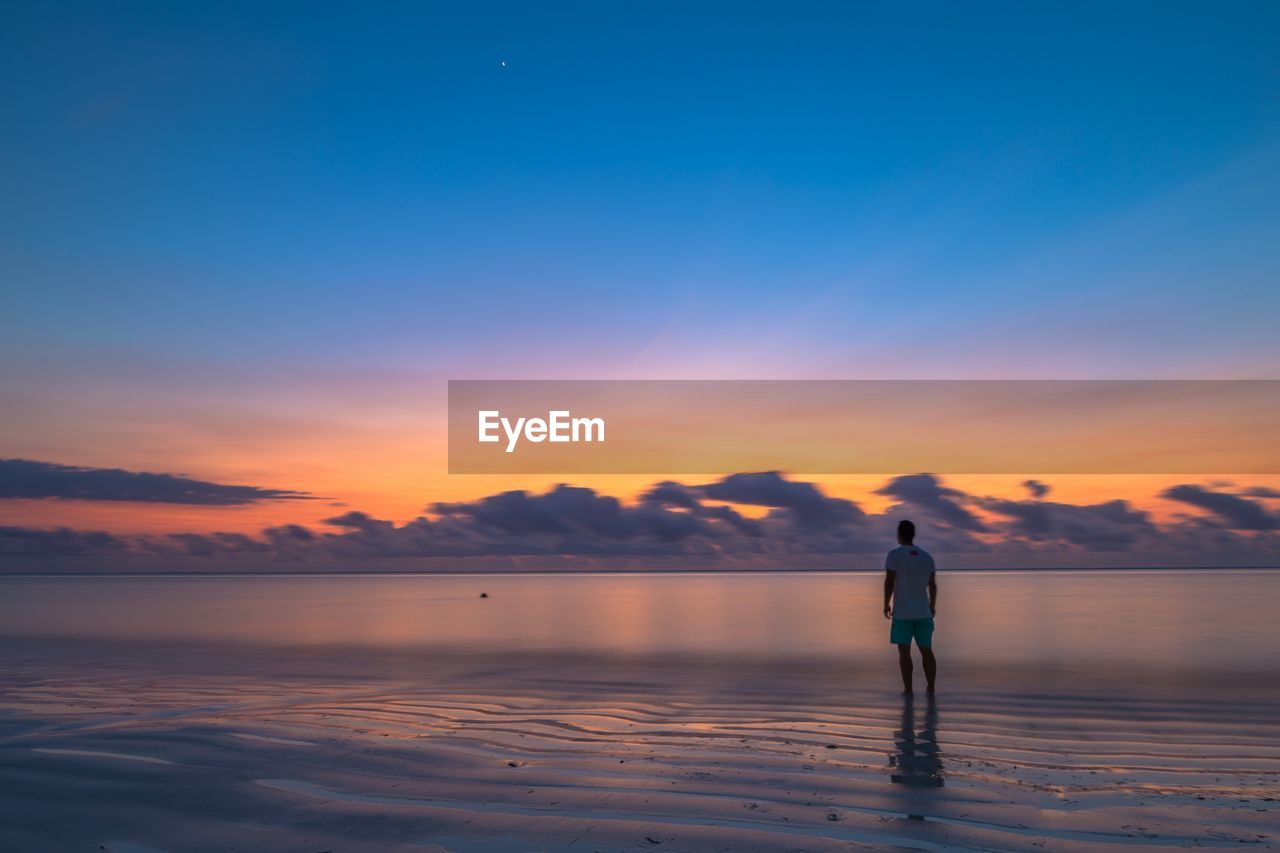 Rear view of man standing on shore at beach against sky during sunset