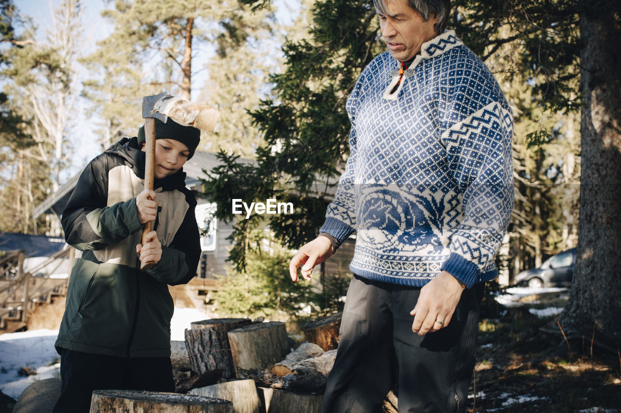 Mature man looking at son cutting log with axe in forest during winter