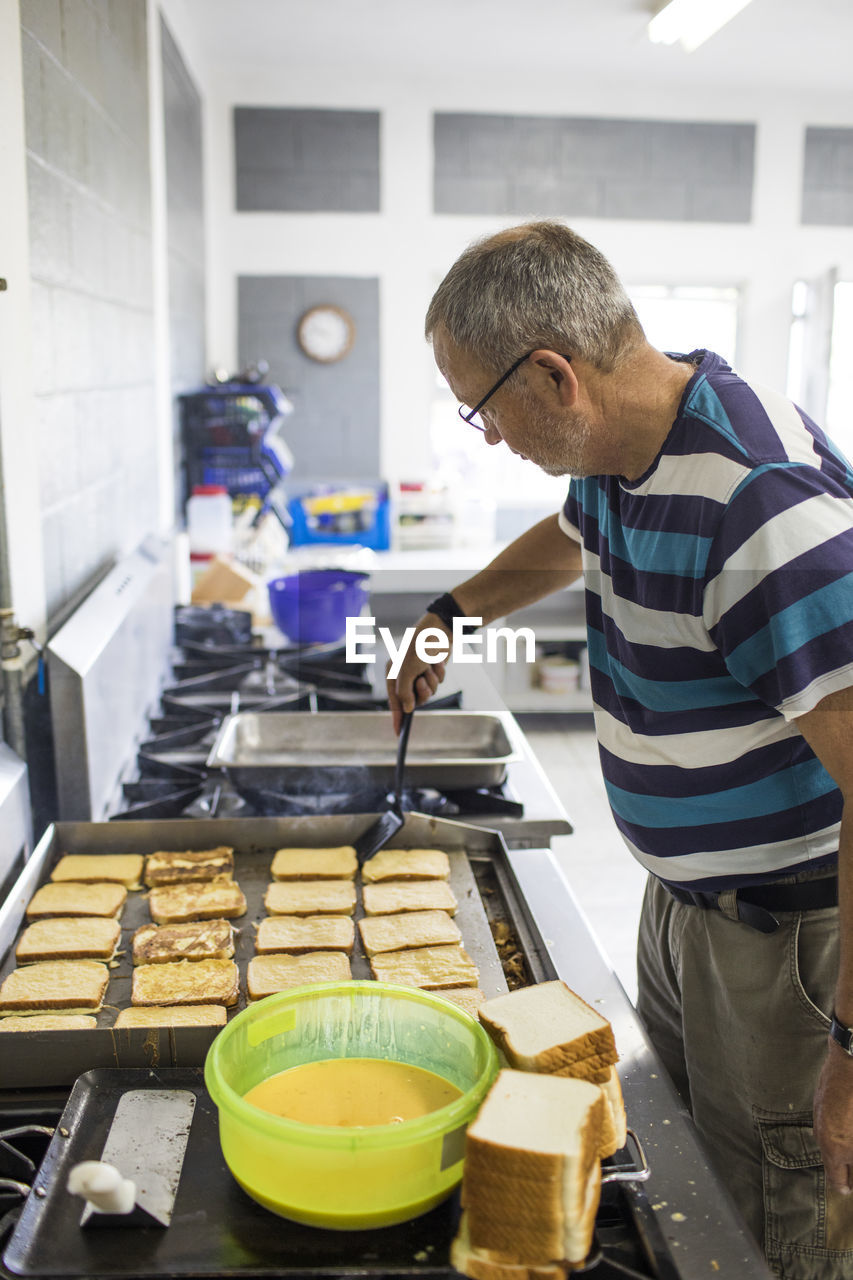 Elderly man cooking french toast in industrial kitchen.