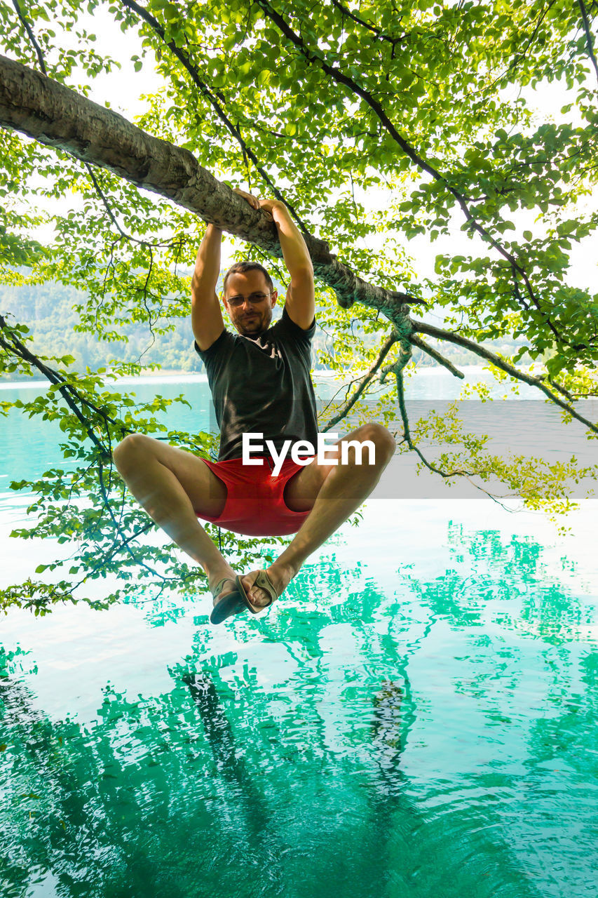 Portrait of young man hanging on branch over lake in forest