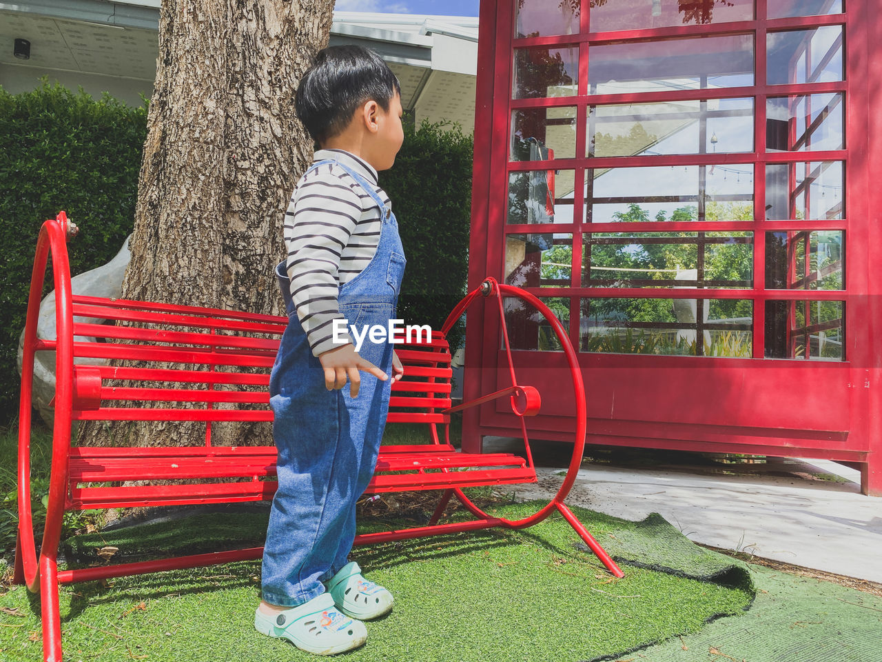 one person, city, full length, outdoor play equipment, public space, playground, childhood, casual clothing, child, men, standing, leisure activity, nature, lifestyles, day, plant, toddler, red, side view, outdoors, architecture, built structure, park, looking, person, adult