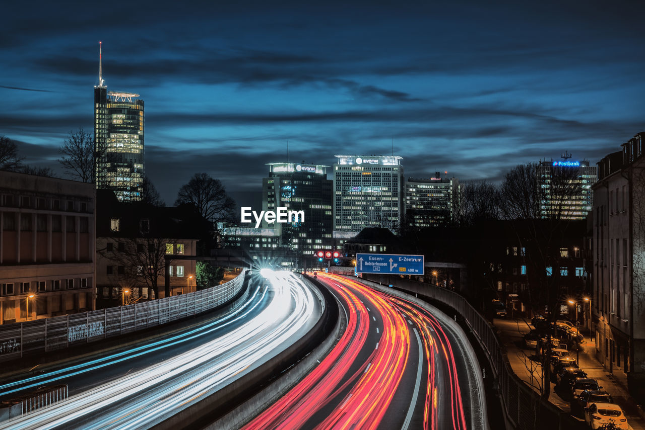 LIGHT TRAILS ON ROAD AMIDST ILLUMINATED BUILDINGS IN CITY AT NIGHT