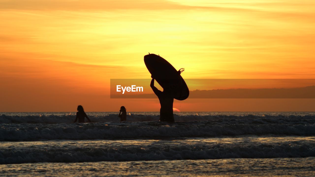 Silhouette surfers standing on beach against sky during sunset