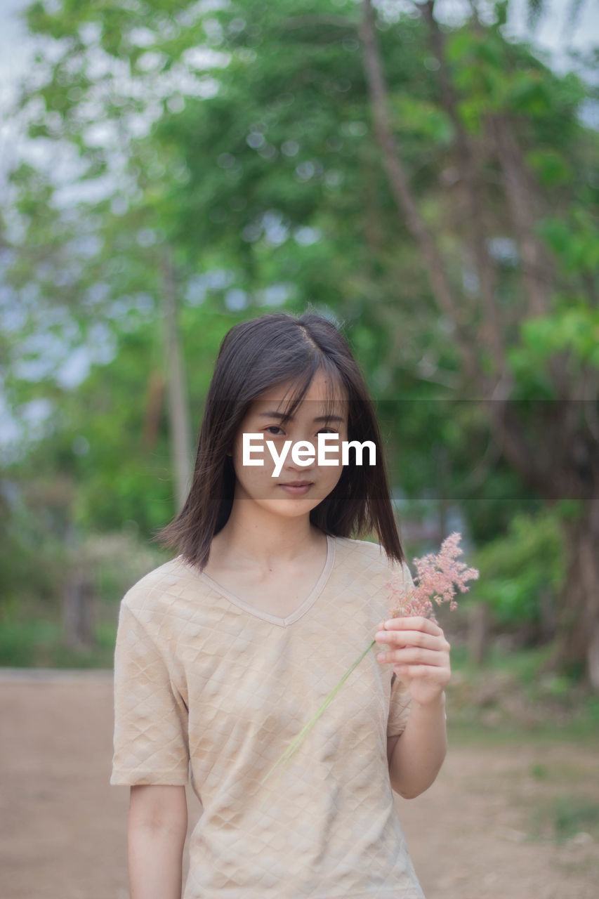 Portrait of beautiful young woman holding flower standing against tree