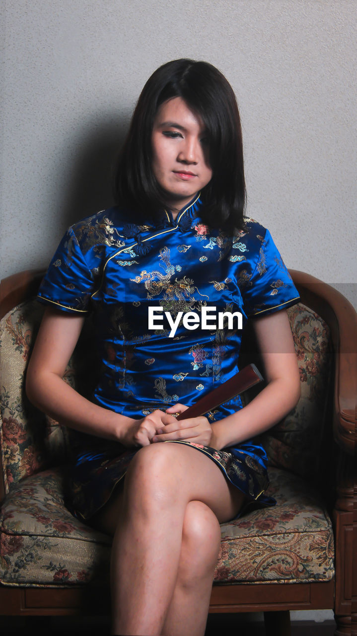 Female model wearing traditional clothing while sitting on chair against wall