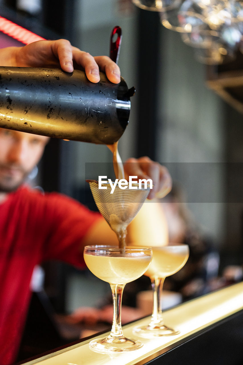 Focused cropped unrecognizable bartender pouring cold refreshing cocktail through strainer in glass placed on counter in bar