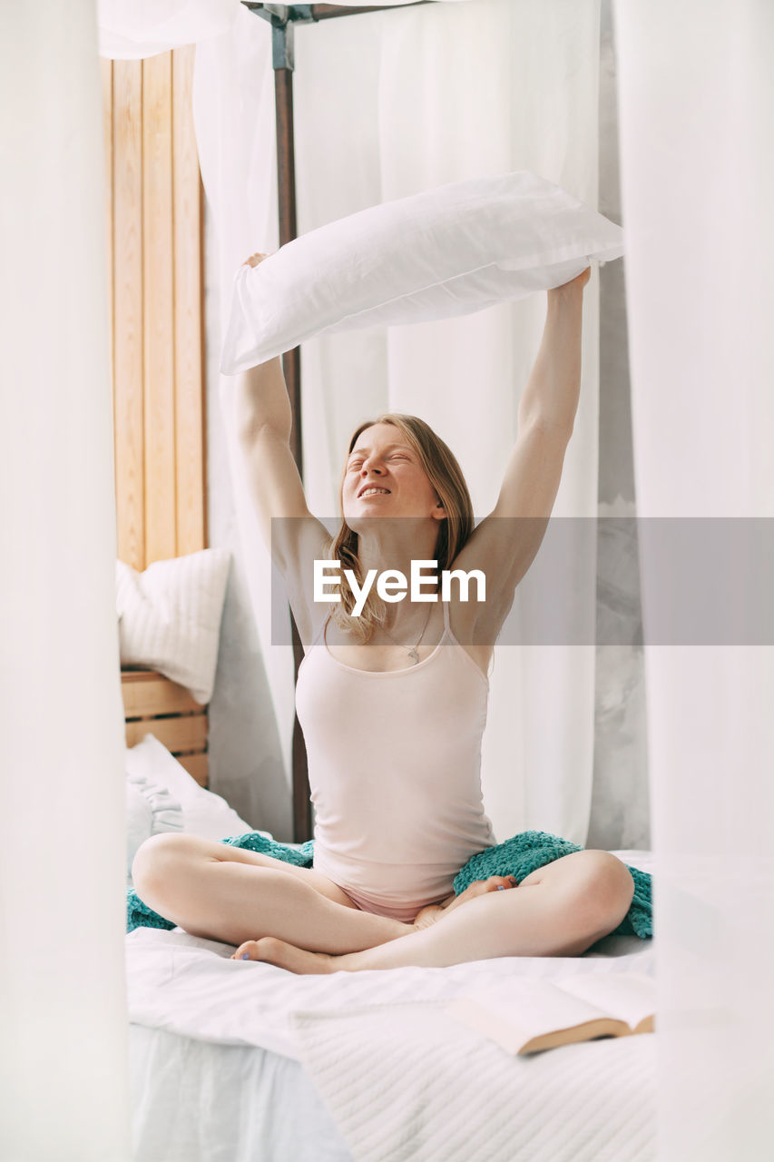 A beautiful smiling girl sits cross-legged on the bed and holds a pillow on her outstretched arms. 