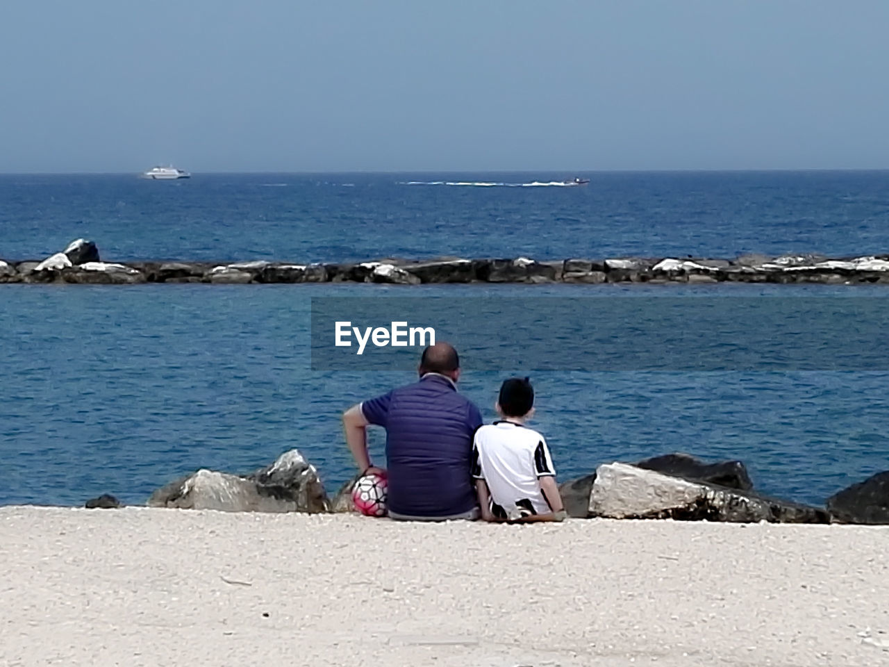 REAR VIEW OF FRIENDS SITTING AT BEACH AGAINST CLEAR SKY