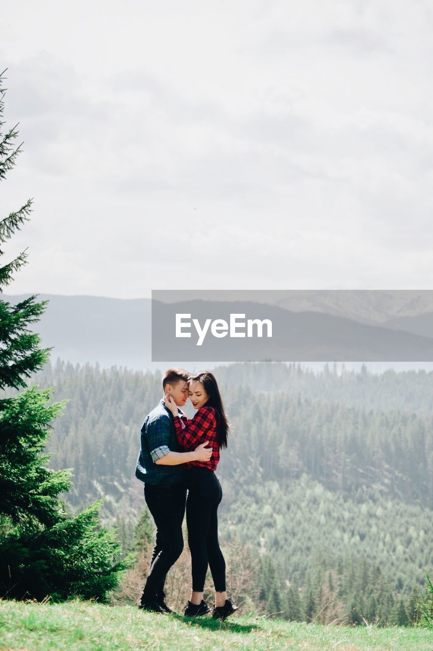 Side view of loving young couple standing against sky in forest during sunny day