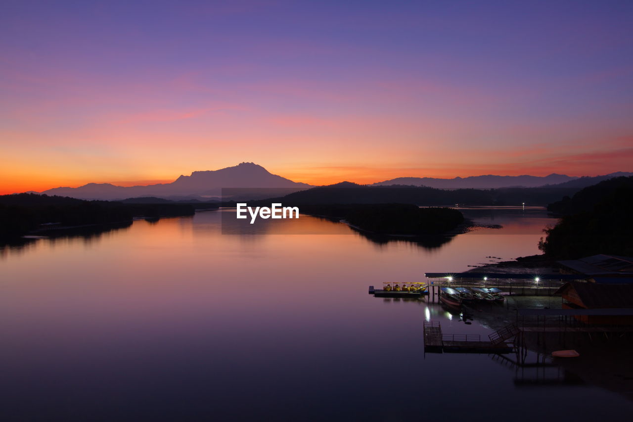 SCENIC VIEW OF LAKE BY SILHOUETTE MOUNTAINS AGAINST SKY AT SUNSET