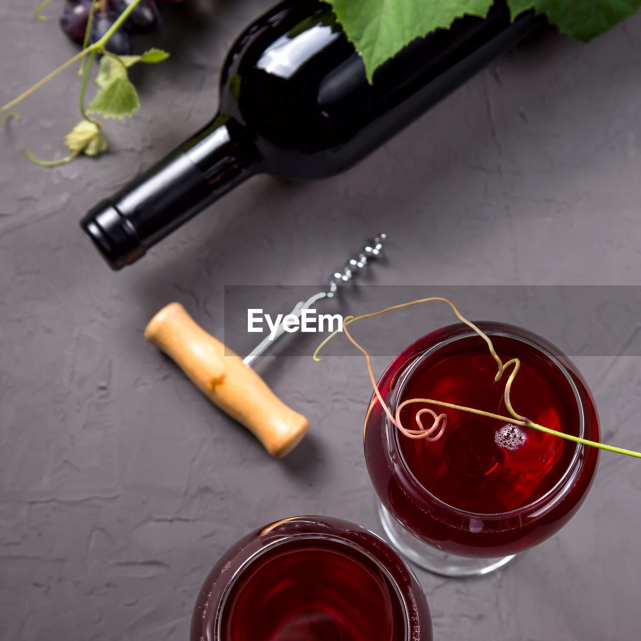 Two glasses of red wine next to a roll, a bunch of grapes and a corkscrew on a gray background