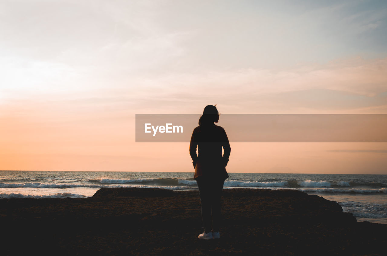 Rear view of silhouette woman standing at beach during sunset