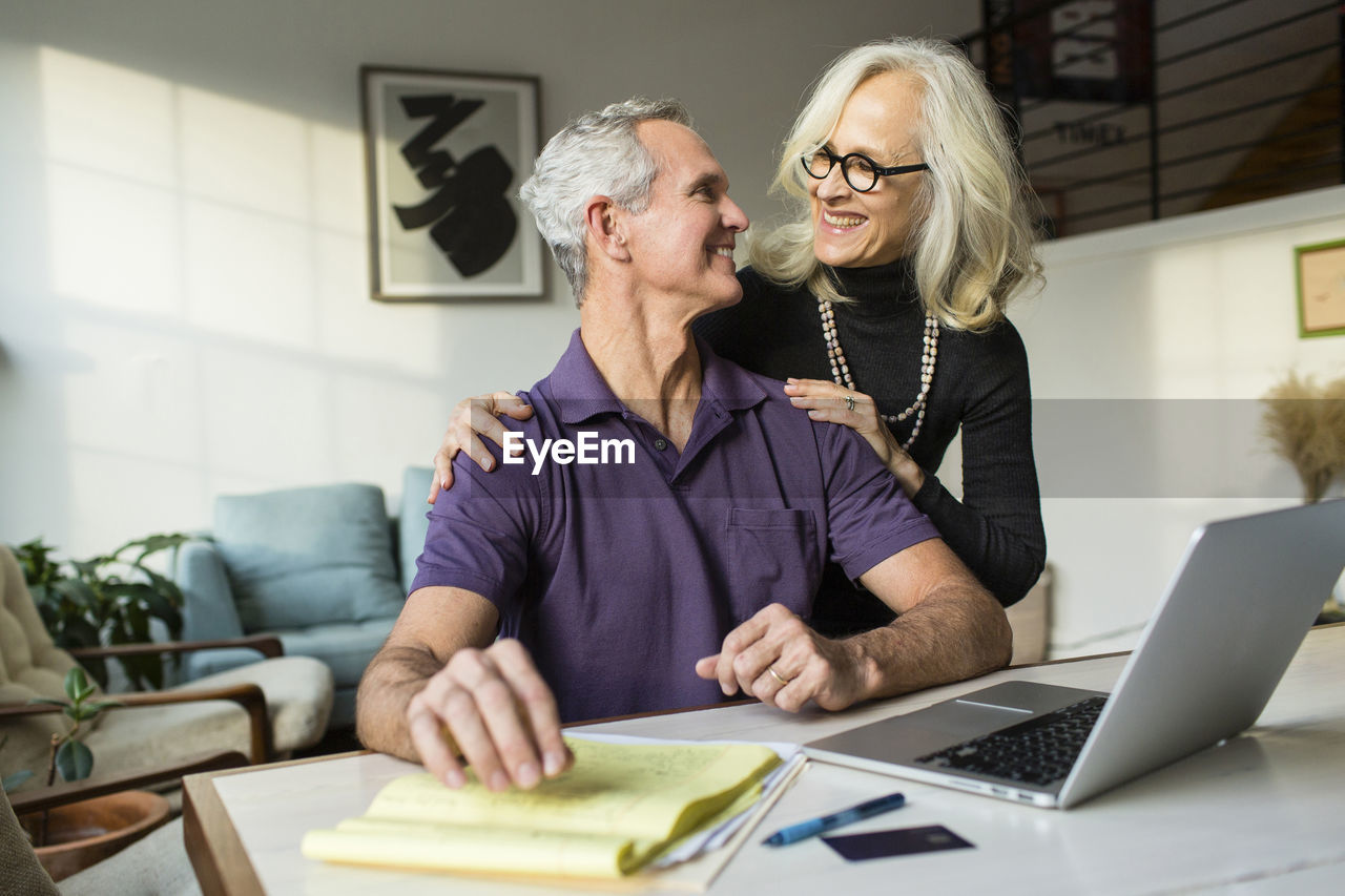 Smiling couple looking each other face to face while using laptop computer at home