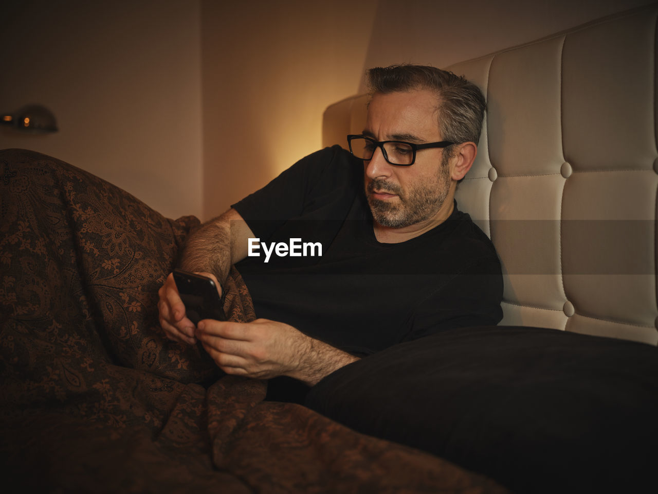 Relaxed and serious man in bed reading on the internet with a smartphone before sleeping and resting