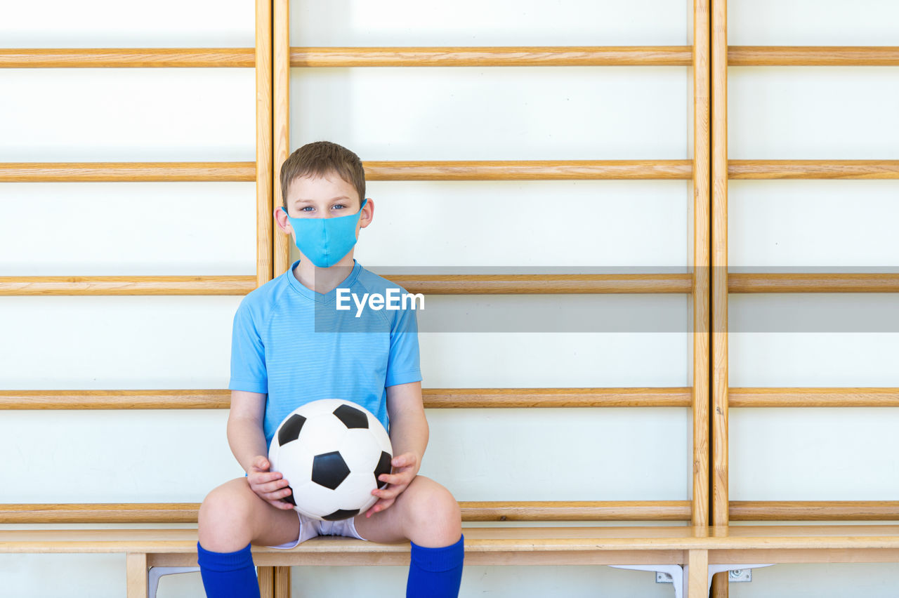 Portrait of boy wearing mask with soccer ball sitting on bench