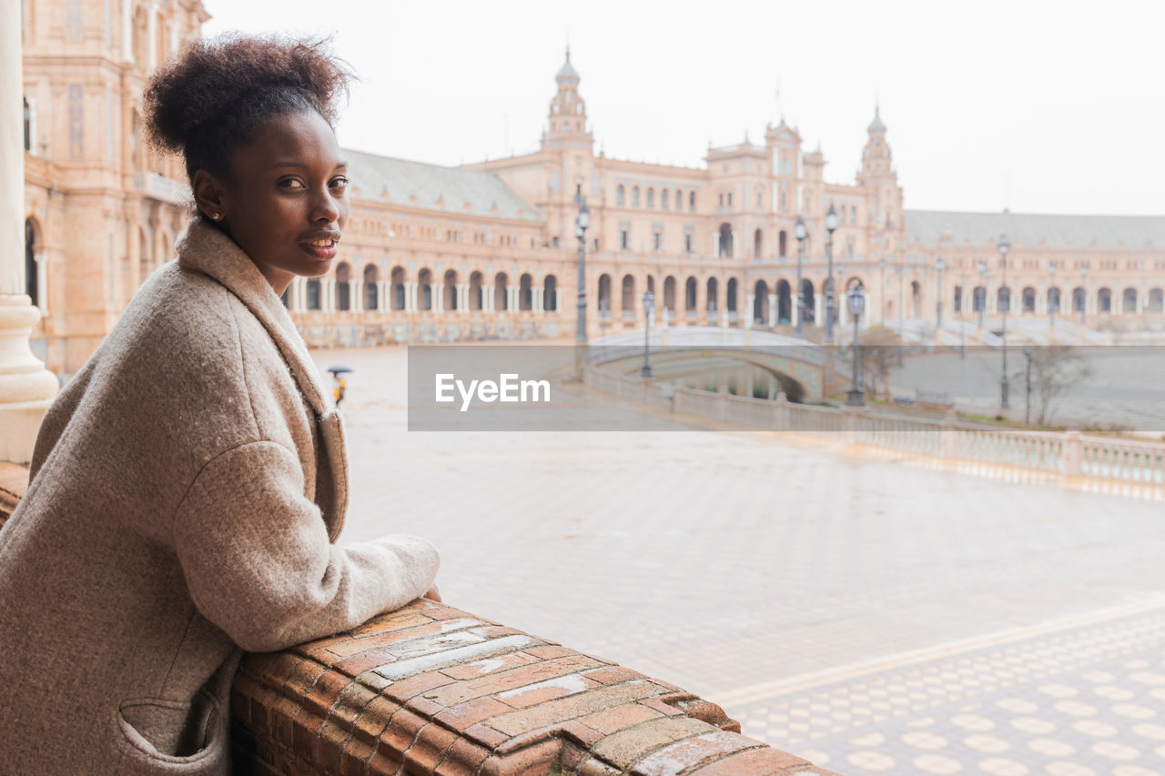Side view of young african american female in casual wear in grand palace with colonnade stucco work ornaments and columns while visiting seville spain while looking at camera