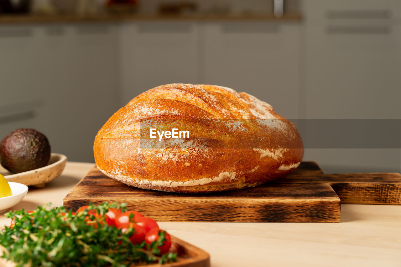 Whole loaf of freshly baked white wheat bread on wooden board on home kitchen table