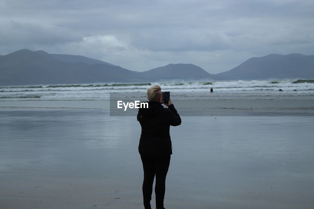 Rear view of woman photographing sea while standing at beach against sky