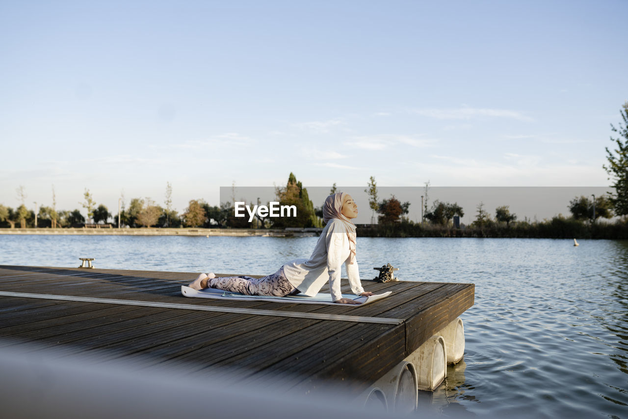 Full body side view of arab female in hijab practicing urdhva mukha shvanasana posture on wooden waterfront near river during yoga session