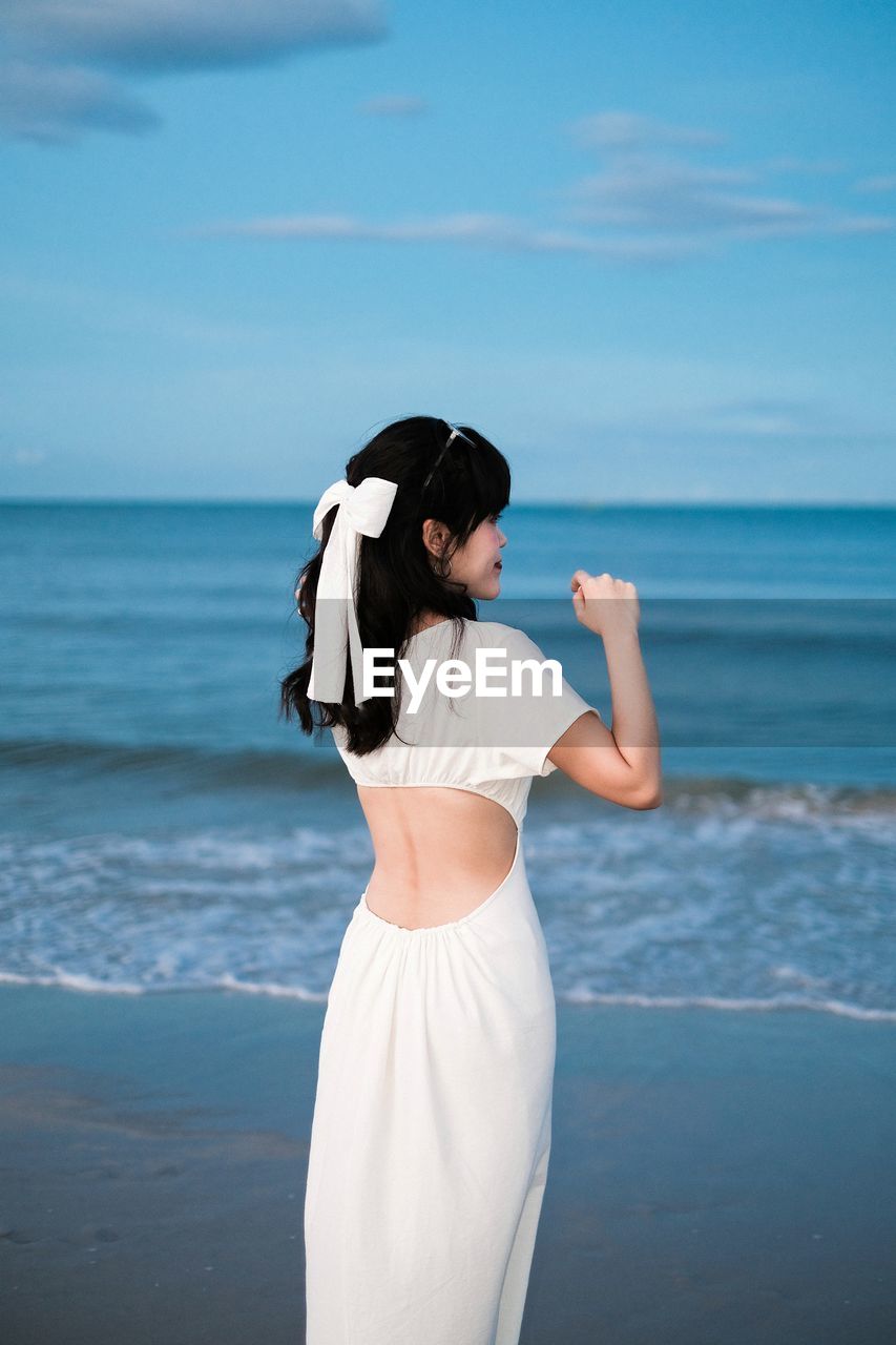 Portrait of young woman wearing white dress standing at beach against sky