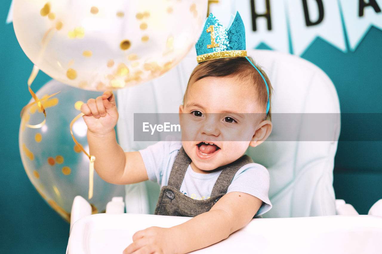 Cute baby boy holding balloon on high chair in birthday party