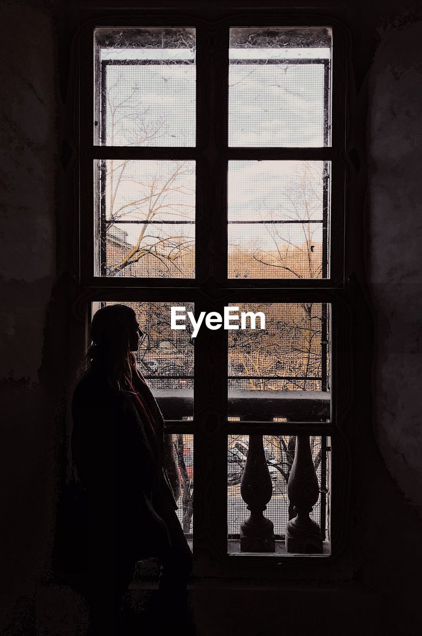 Silhouette mid adult woman looking through window while standing at home