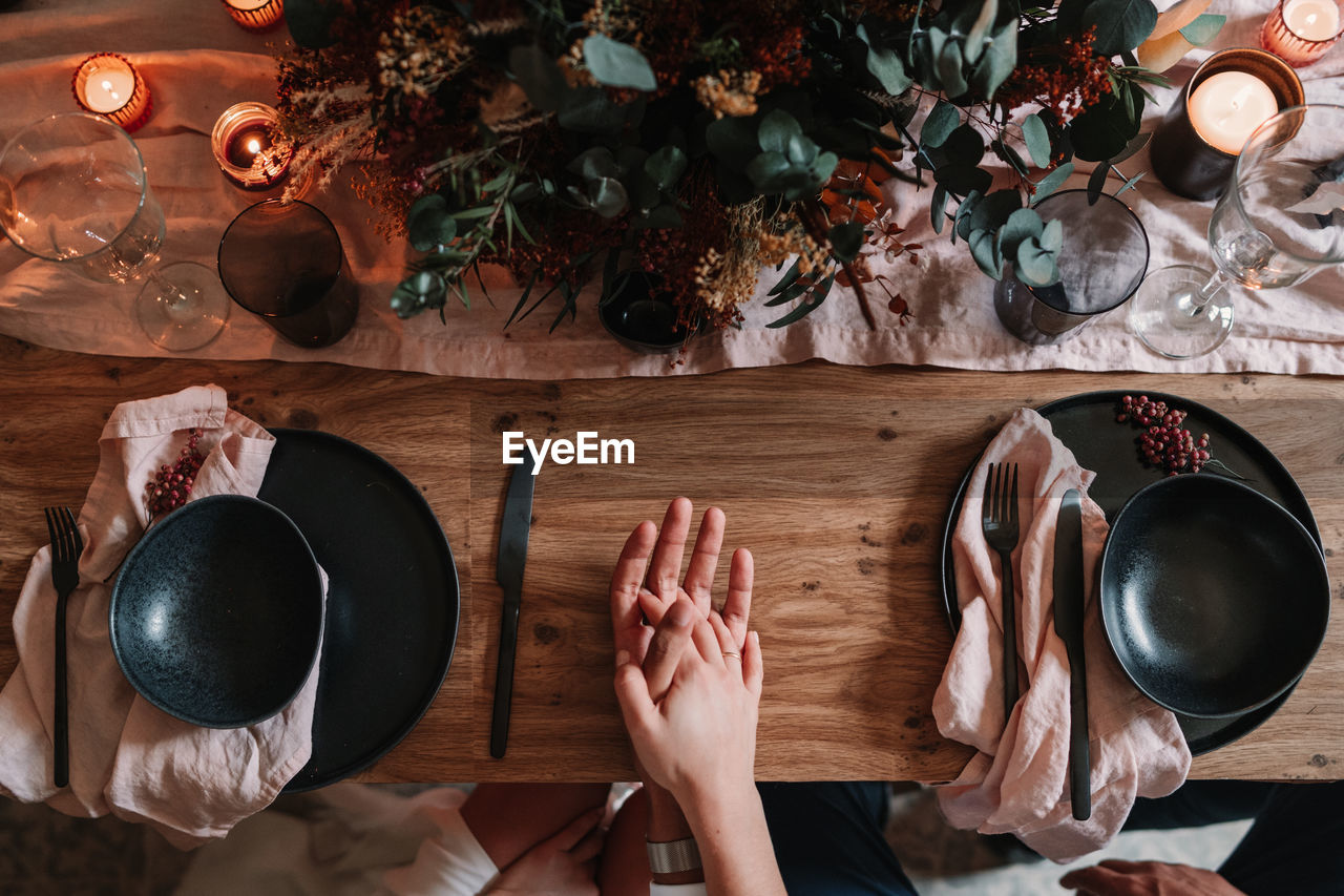 Top view of crop anonymous groom and bride holding hands on served table with flower decor and burning candles in restaurant