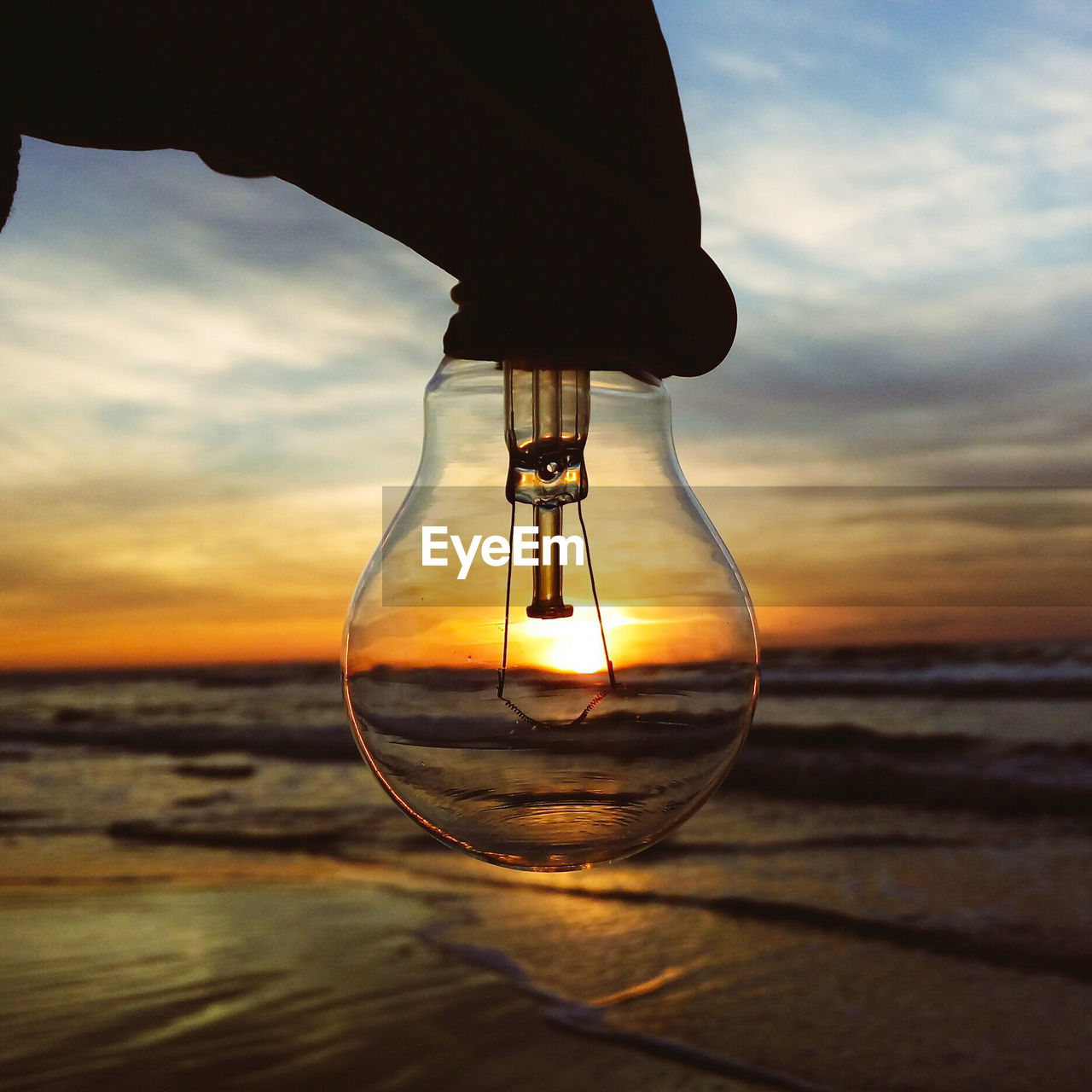 Cropped silhouette hand of person holding light bulb at beach against sky during sunset