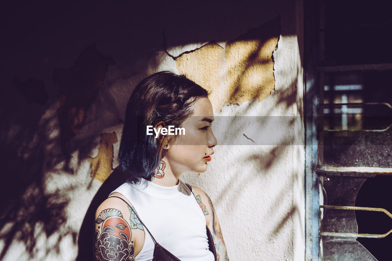 Thoughtful young woman with tattoos standing by old house