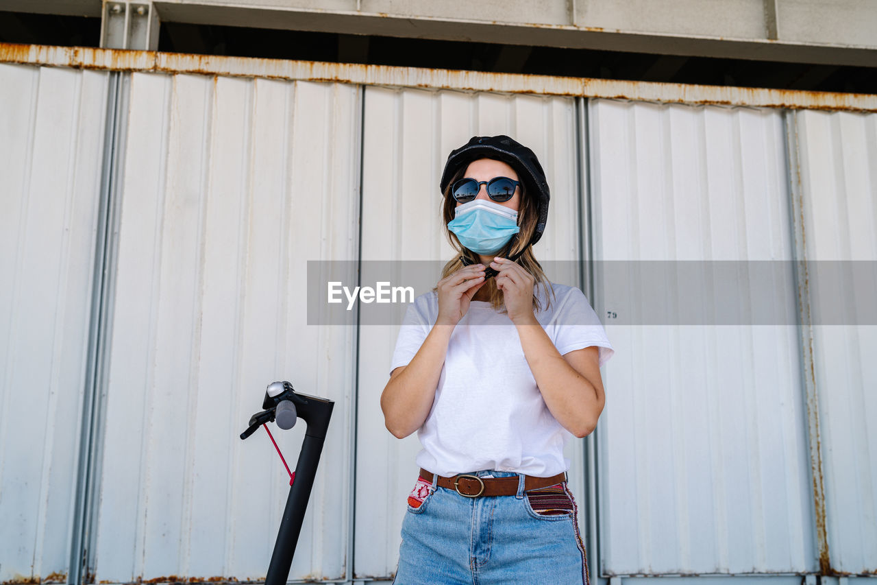 Young female rider in medical mask and sunglasses putting on protective helmet while preparing for riding electric scooter