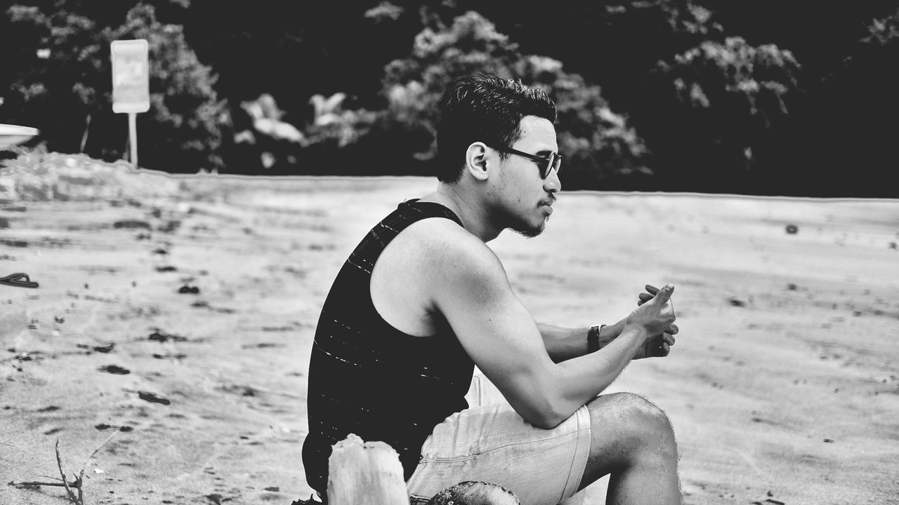 Side view of young man wearing sunglasses sitting at beach
