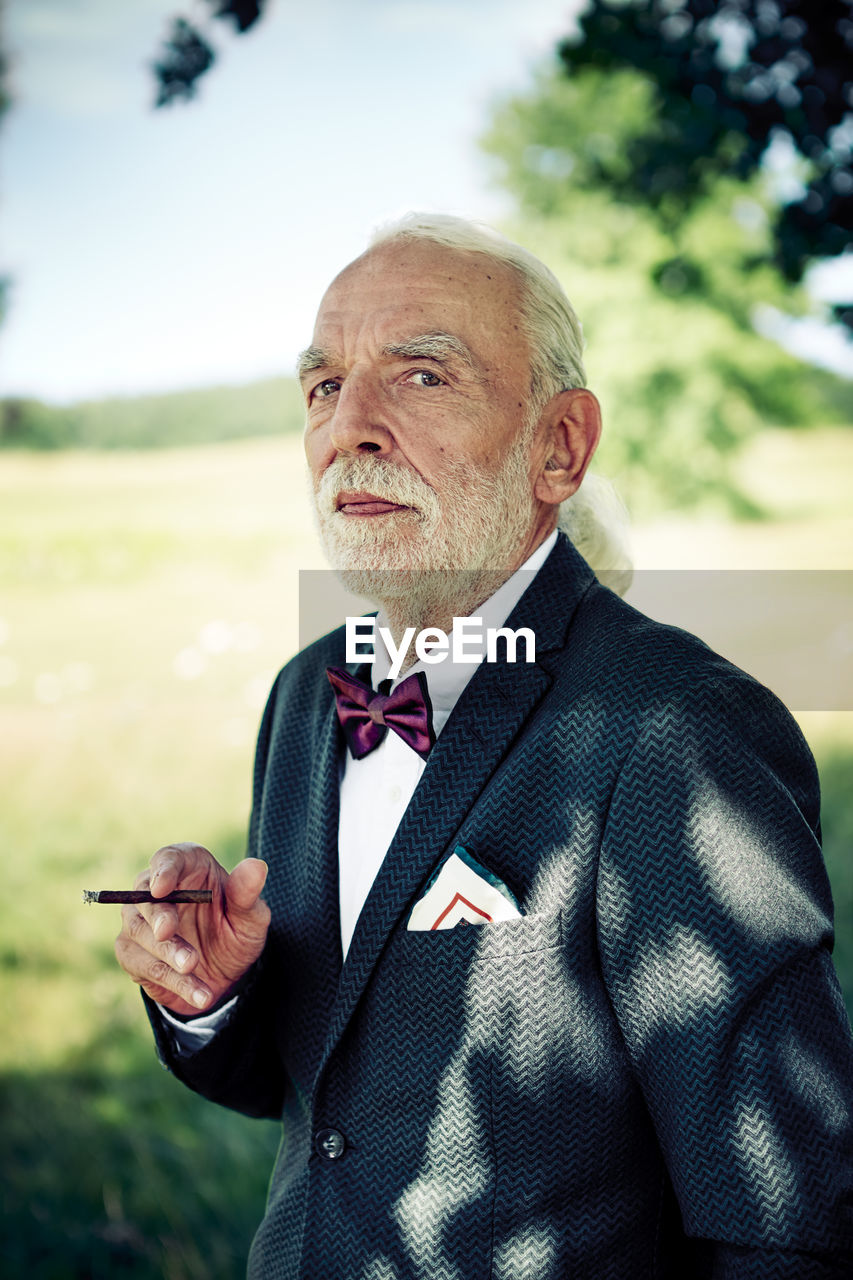 Portrait of senior man wearing suit coat, bow tie and dress handkerchief in nature smoking cigarillo