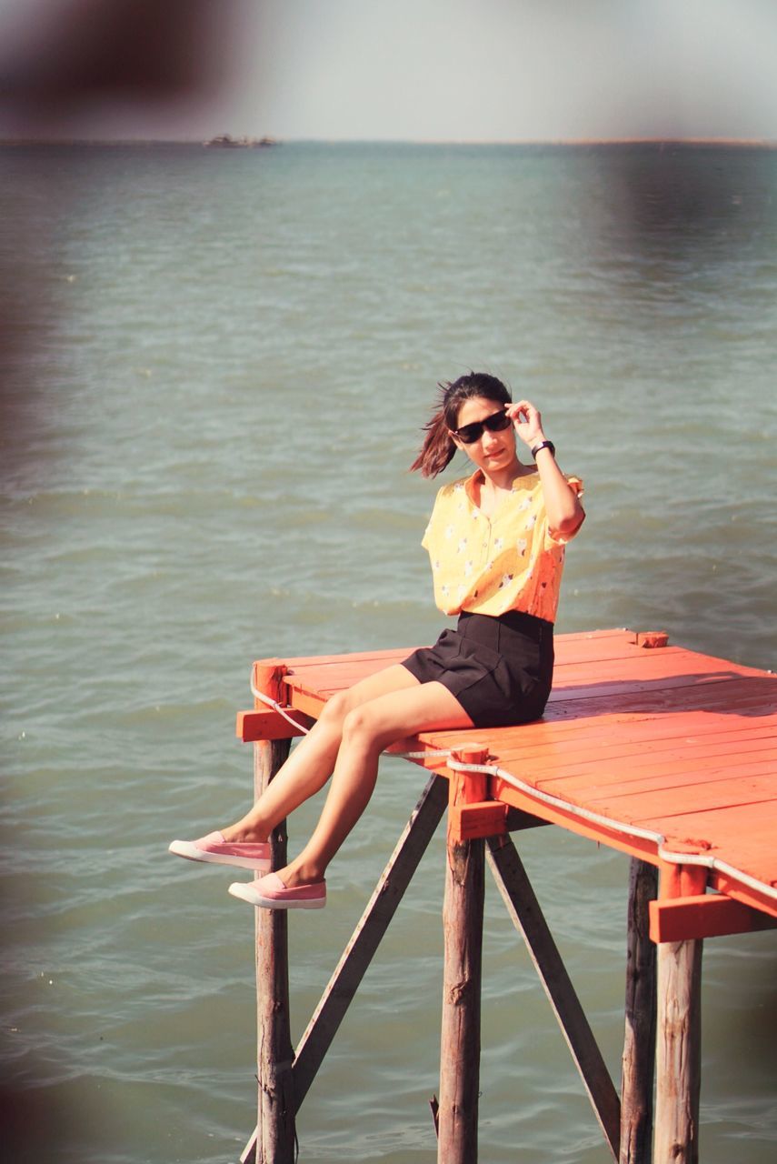 FULL LENGTH OF YOUNG WOMAN SITTING ON RAILING AGAINST SEA