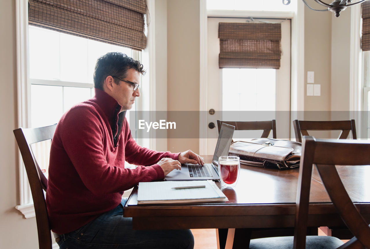 Man in glasses working from home using a computer at a dining table.