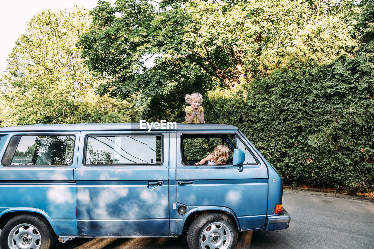 Two girls sitting on and in vintage blue van in summertime