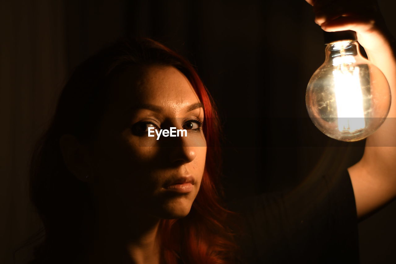 Close-up portrait of young woman holding light bulb on black background