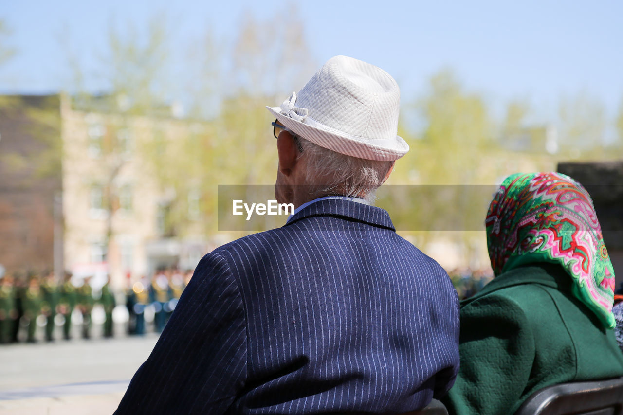 REAR VIEW OF MAN WITH HAT SITTING ON OUTDOORS