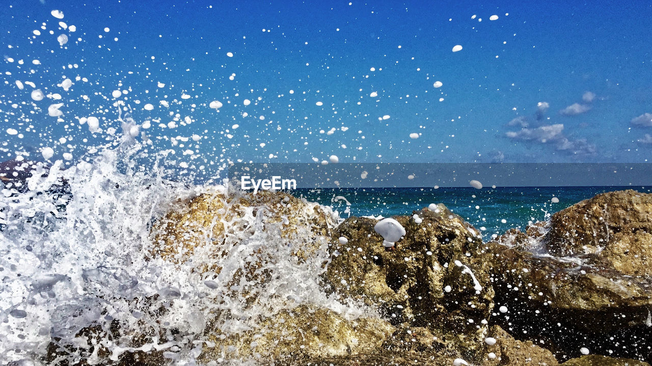 Water splashing on rocks at beach against blue sky