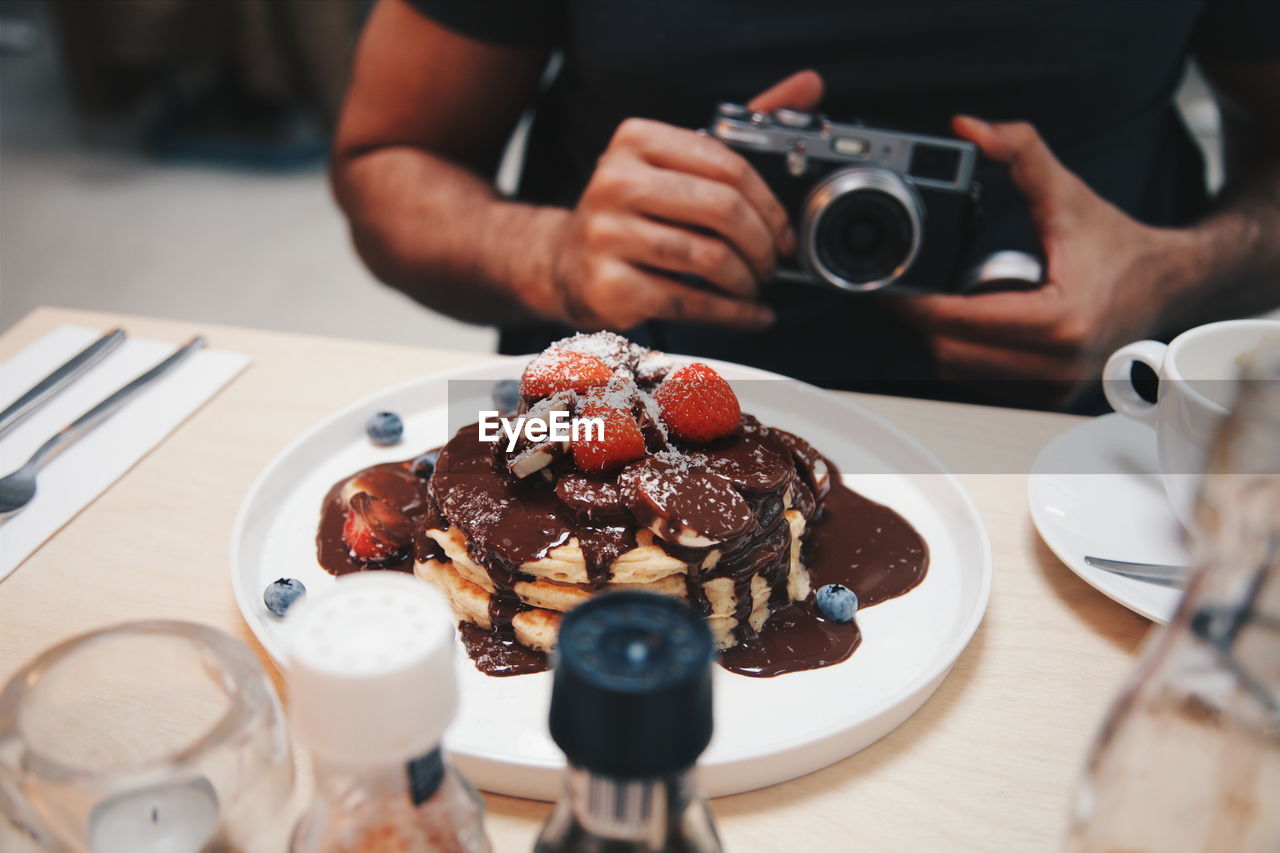 Midsection of man with camera sitting by pancake in plate on table at restaurant 