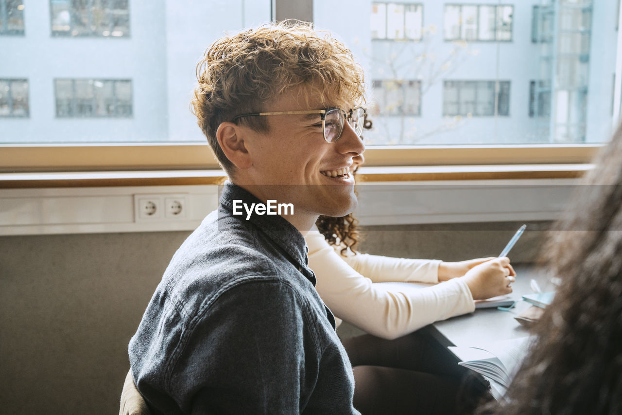 Side view of smiling young blond man wearing eyeglasses sitting in classroom